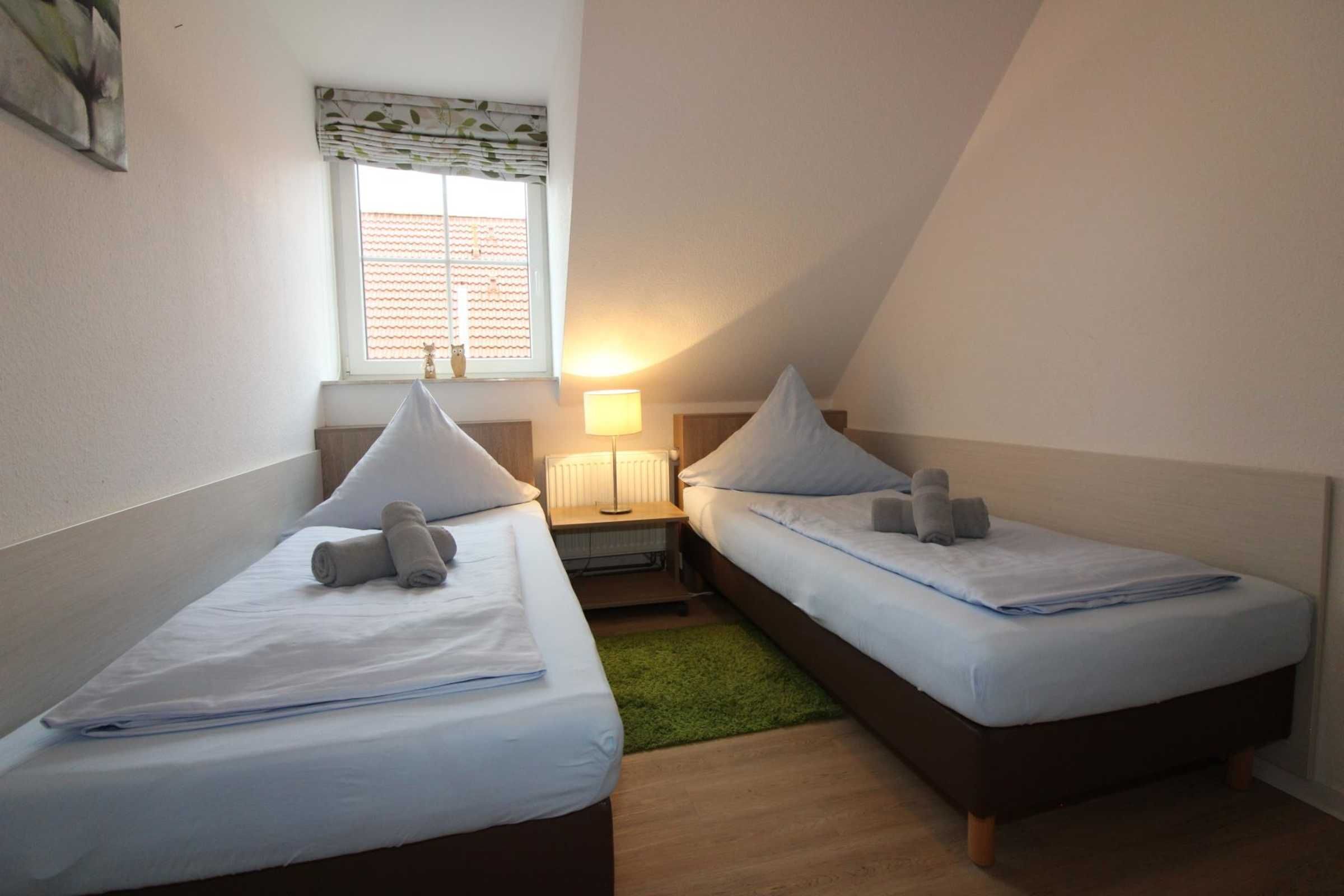 Une petite chambre agréable, dotée de deux lits simples, d’une table de chevet avec une lampe, d’une fenêtre avec des stores et d’un tapis vert.