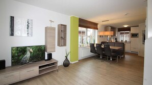 A modern living room with a flat-screen TV, a dining area with a table and chairs, and a kitchen in the background. - ROB 17 for 6 persons, approx. 86 sqm, pets - Feriendorf Robbenplatte Burhave (Butjadingen)