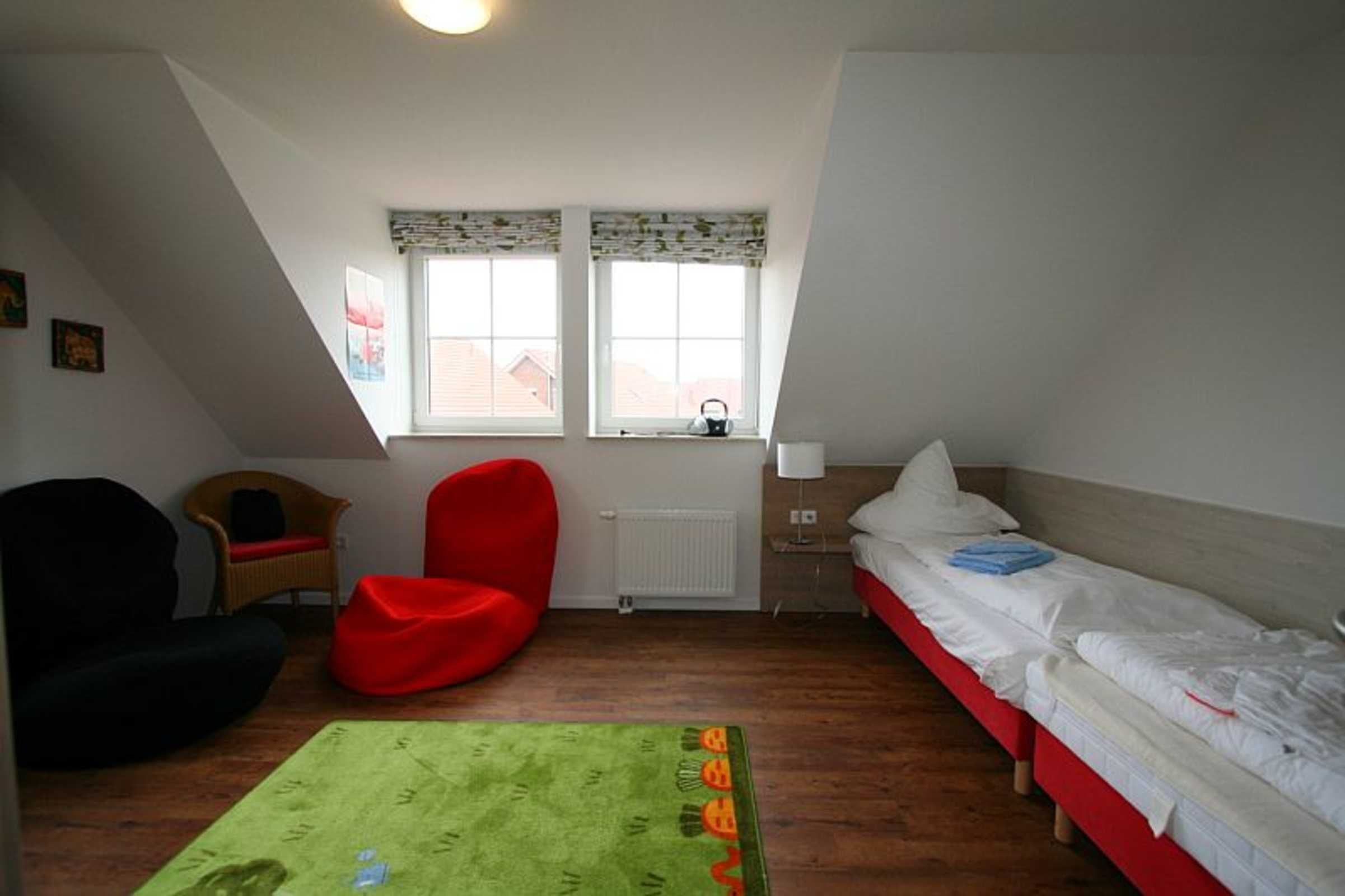 A small, well-lit room with a sloped ceiling, a single bed, a red chair, a small table, and a window with a view of buildings outside.