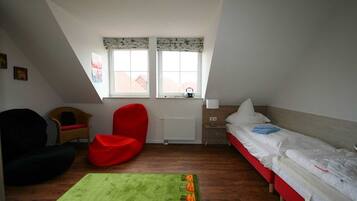 A small, well-lit room with a sloped ceiling, a single bed, a red chair, a small table, and a window with a view of buildings outside.
