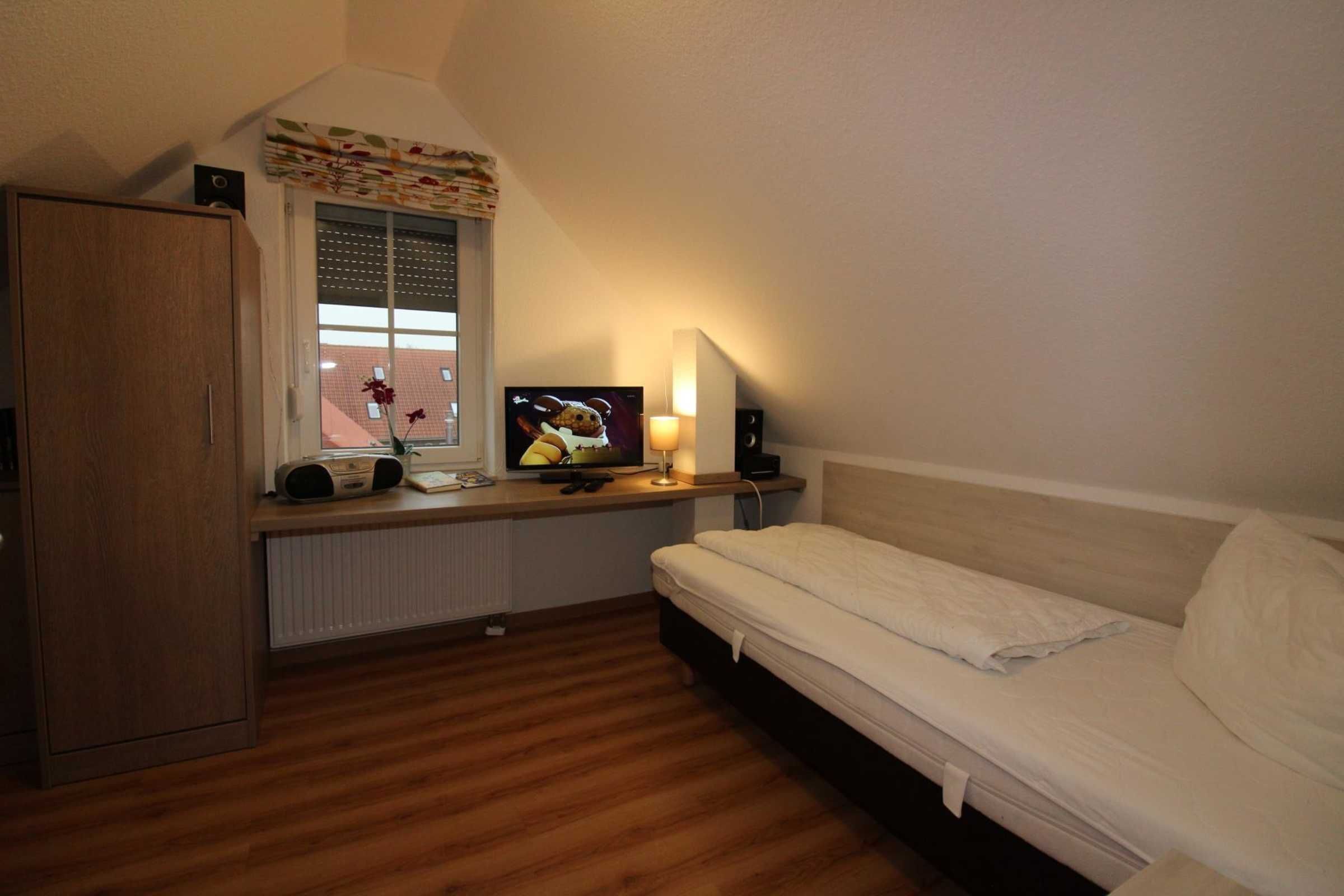 A bedroom with a wooden floor, a bed, a desk with a computer and a lamp, a window with a view of buildings, and a radiator.