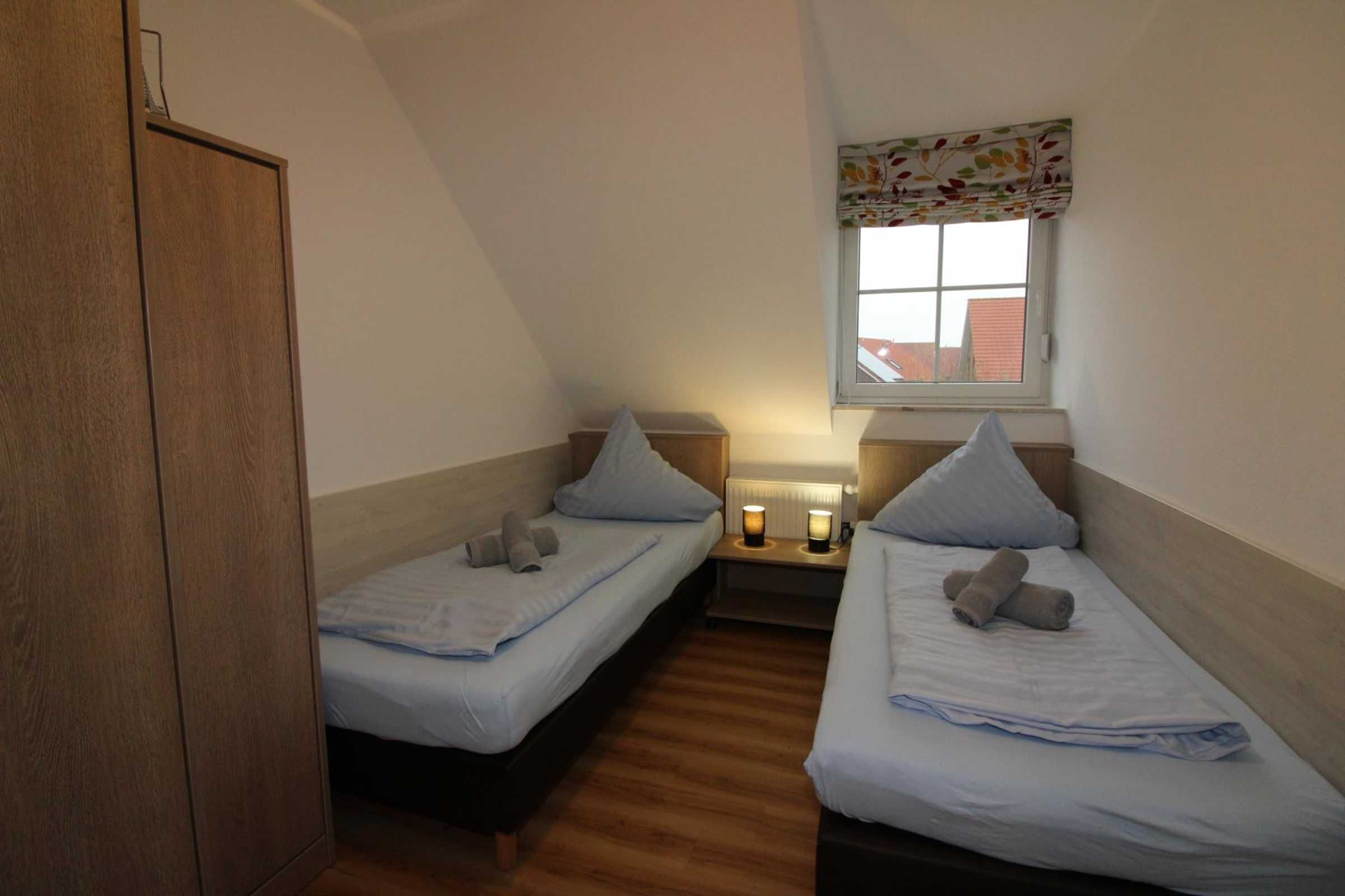 Una habitación pequeña y acogedora con dos camas individuales, suelo de madera, una ventana con un adornado floral y una mesita de noche con lámpara.