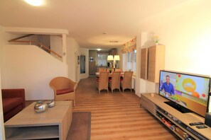 A living room with a TV, a dining area, and a kitchen in the background.