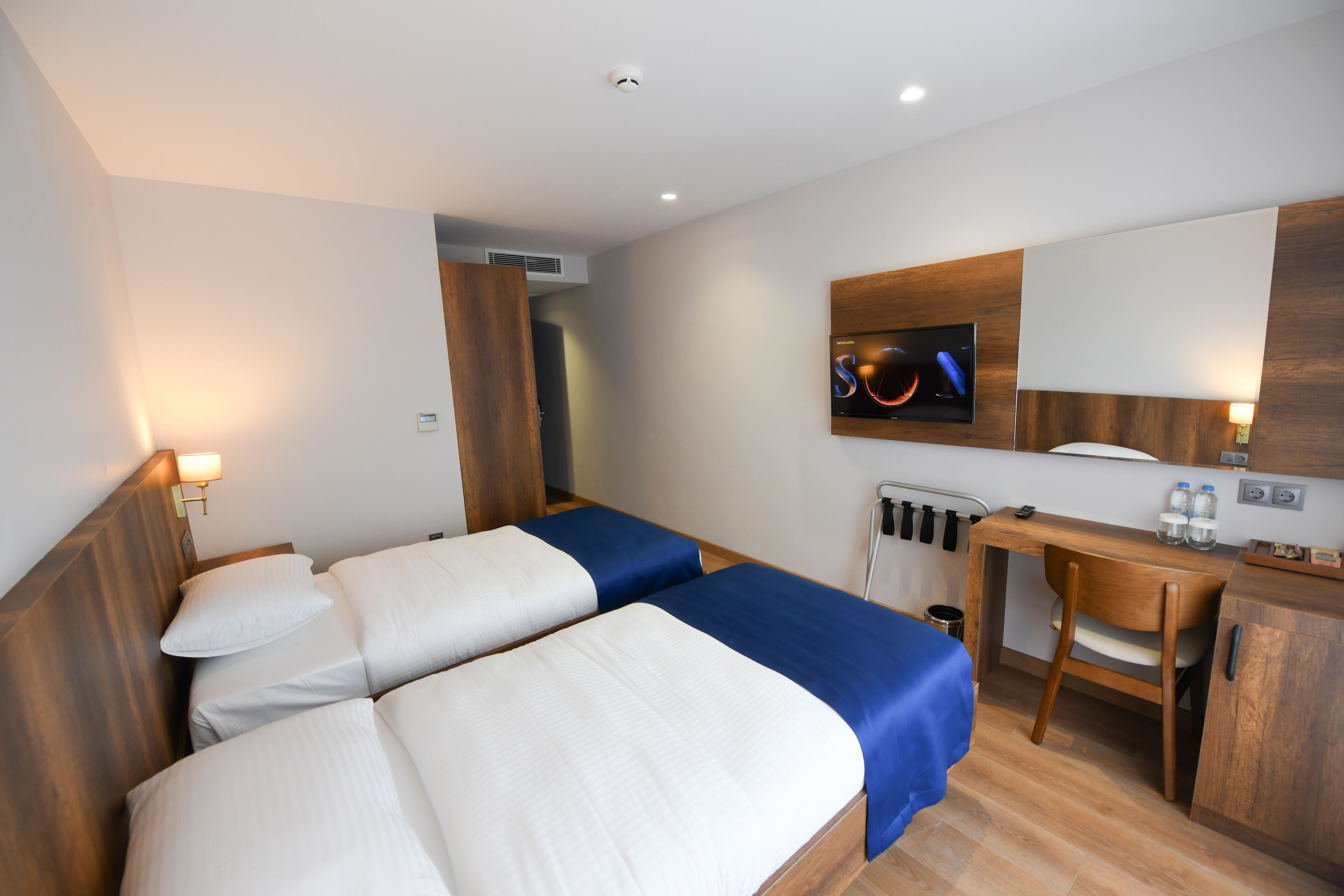 A hotel room with two beds, a desk, a chair, a TV, and a small kitchenette.
