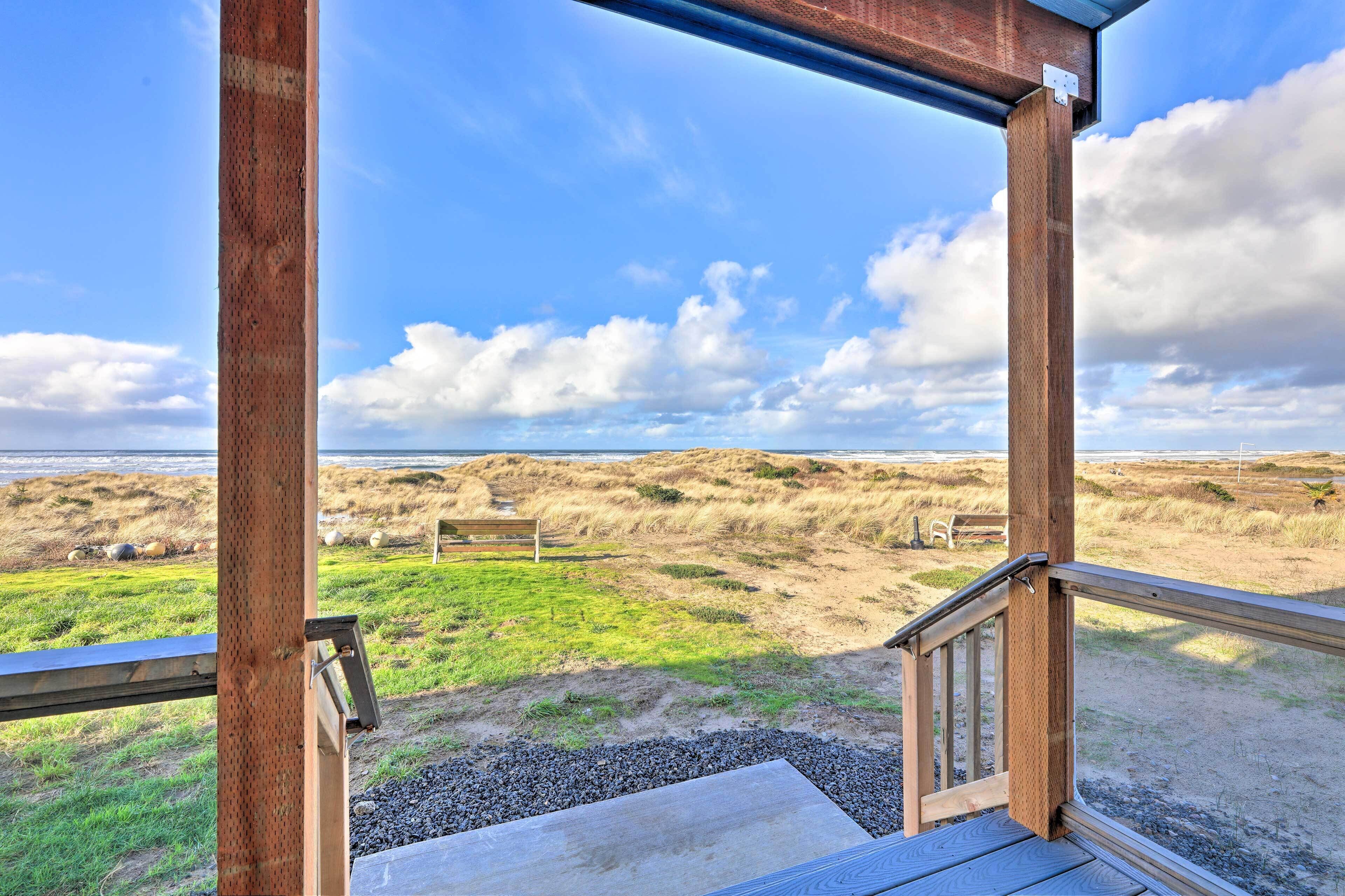 Une terrasse en bois avec une rambarde en métal, donnant sur une plage bordée de dunes et agrémentée d’un banc.