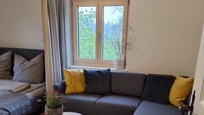 A living room with a grey sectional sofa, a round wooden coffee table, a potted plant, and a window with a view of greenery. - Spacious vacation apartment for 2 to 4 people surrounded by nature (Feldkirch)