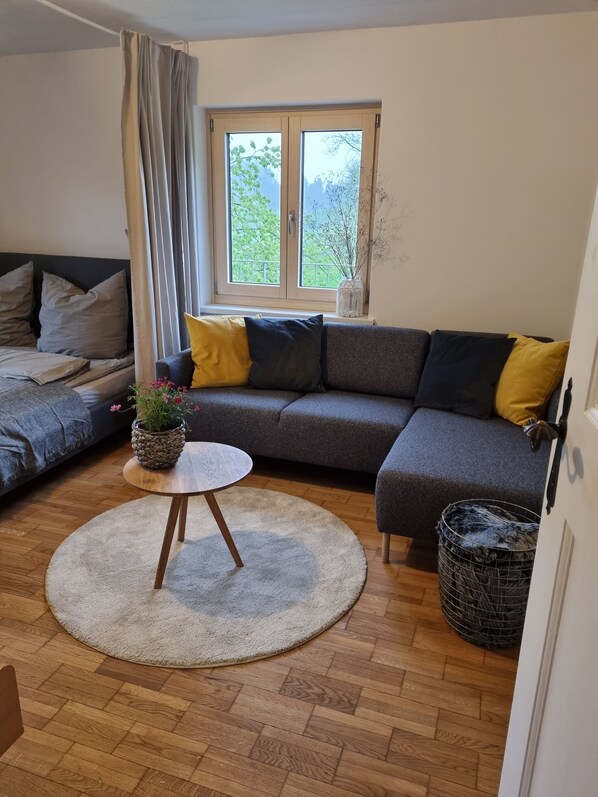 A living room with a grey sectional sofa, a round wooden coffee table, a potted plant, and a window with a view of greenery.