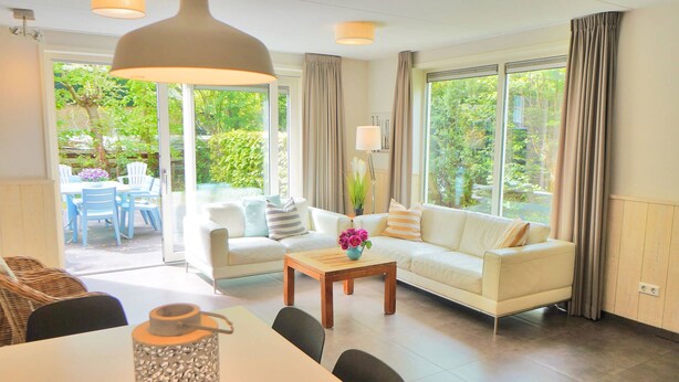 Un salon spacieux comprenant un canapé, une table basse et un coin repas. De grandes fenêtres laissent entrer la lumière naturelle et offrent une vue sur la verdure environnante.