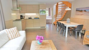 Une cuisine moderne avec des meubles en bois, un coin repas avec une table et des chaises blanches, un escalier et un salon avec un canapé et une table basse.