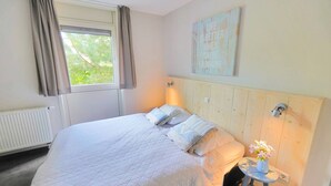 Une chambre avec un grand lit, une fenêtre avec des rideaux, une lampe fixée au mur et une petite table sur laquelle est posé un vase de fleurs.