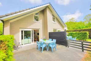 Ein modernes Haus mit einer Terrasse, einem Tisch, Stühlen, einer Glastür und einem eingezäunten Garten.
