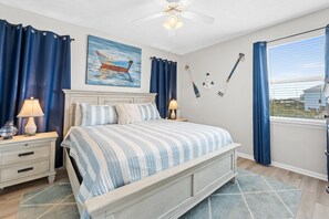 Ein Schlafzimmer mit einem Bett, zwei Nachttischen, einem Deckenventilator, einem Gemälde eines Bootes und einem Fenster mit Vorhängen.