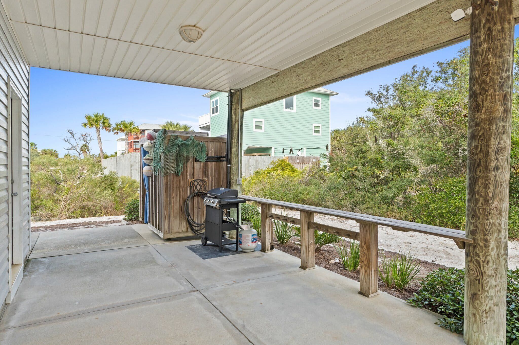 A covered patio with a grill and propane tank, a wooden bench, and a view of a green house and trees.