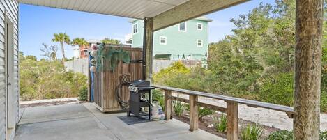 A covered patio with a grill and propane tank, a wooden bench, and a view of a green house and trees.