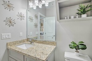 Un baño con lavamanos de encimera de granito, inodoro blanco, una planta en maceta y decoraciones con forma de estrella en la pared.