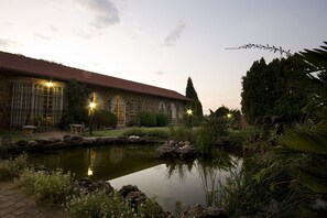 A brick building with arched windows and a red-tiled roof, surrounded by a pond and greenery. - Farmers Folly Guest House (Pretoria)
