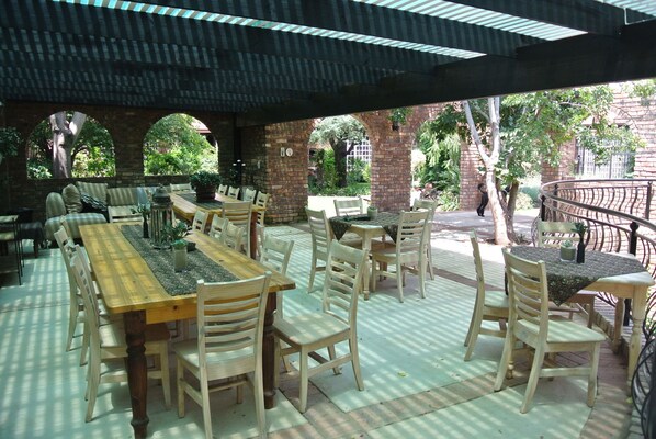 An outdoor patio with multiple wooden tables and chairs, brick walls, and a metal railing. - Farmers Folly Guest House (Pretoria)