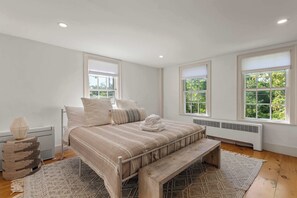 Ein Schlafzimmer mit Bett, Kissen, einem Nachttisch, einer Lampe, einer Heizung und großen Fenstern mit Blick auf Grünflächen.