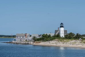 A lighthouse with a white tower and black top, situated on a sandy beach with a wooden pier extending into calm water.