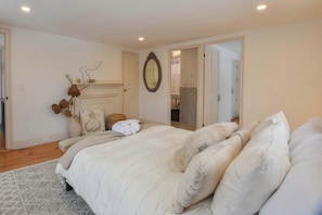 A bedroom with a bed, pillows, a mirror, a fireplace, and a door leading to another room.