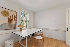 A room with a white table, two wooden chairs, a lamp, a trash bin, and a window with a view of trees. - Mins to Beach, NEW AC and Game Room in 2026! (Barnstable)