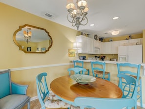 A dining area with a wooden table and blue chairs, a chandelier, and white kitchen cabinets in the background. - Ocean Views, Balcony Swing, Heated Pool Access (Tybee Island)