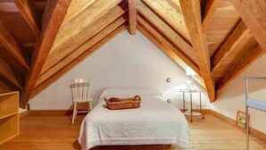 A bedroom with a sloped ceiling, wooden beams, a bed with a white bedspread, a wicker basket, a white chair, and a bedside table with a lamp.
