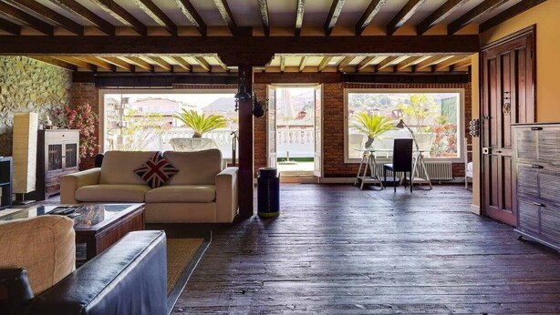 A spacious room with a wooden floor, a sofa, a table, and a chair. There is a large window providing a view of the outside.