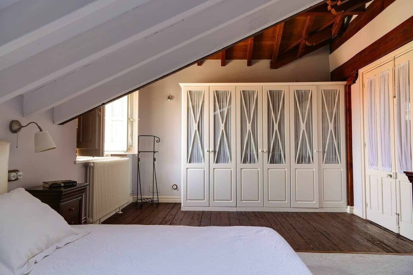 Een slaapkamer met een schuine kap, een bed, een radiator, een kledingkast en een raam.