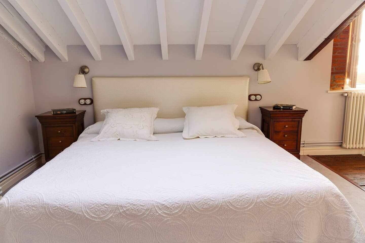 A bedroom with a large bed, two bedside tables, a headboard, and a window with a radiator.
