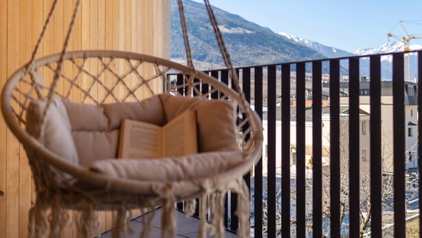 Ein Korbstuhl auf einem Balkon mit Blick auf Berge und Gebäude.