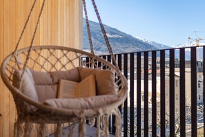 Une chaise en osier sur un balcon, avec vue sur les montagnes et les bâtiments.