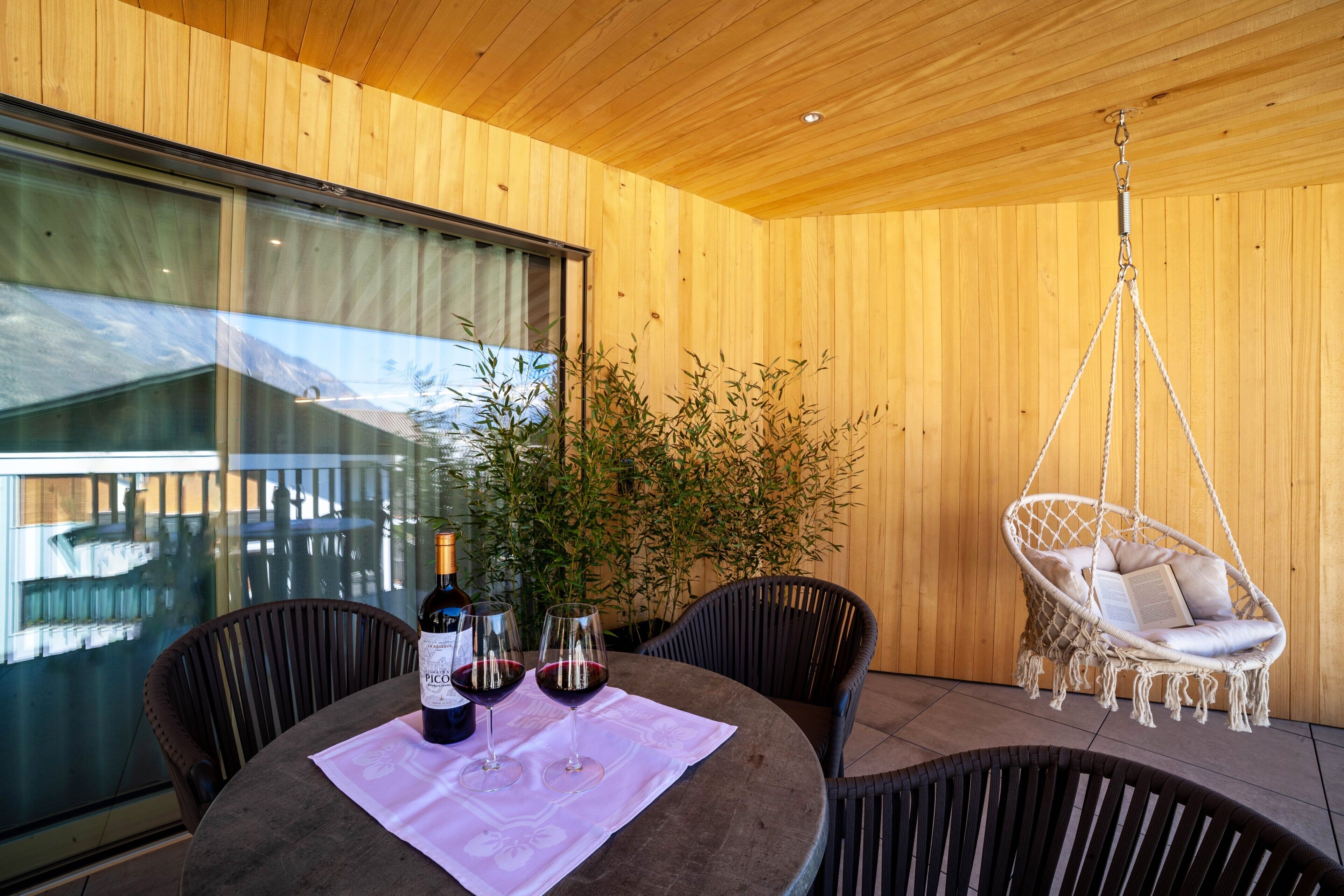 A wooden interior with a round table, two chairs, a hanging chair, and a large window.
