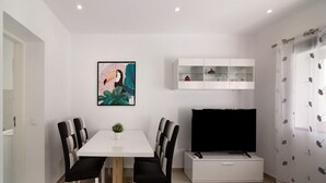 Een moderne woonkamer met een witte eettafel, zwart-wit gestreepte stoelen, een flatscreen televisie, een witte kast en een ingelijst schilderij aan de muur.