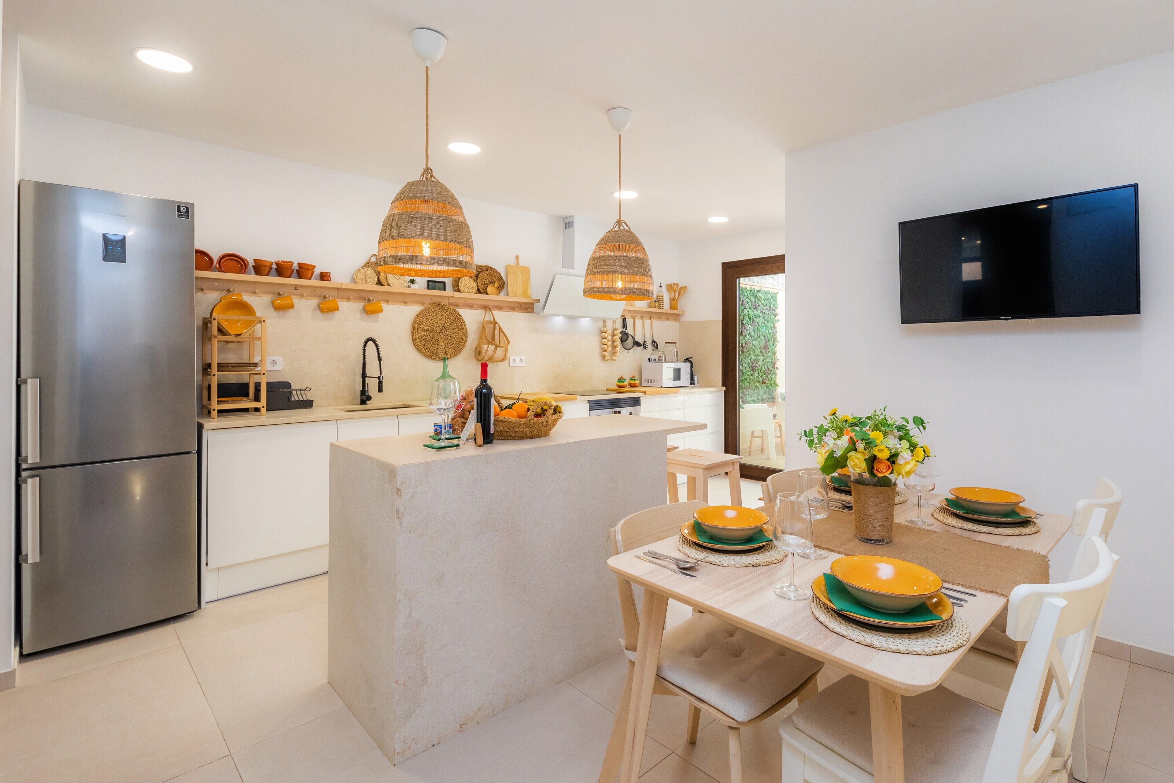 A modern kitchen with a central island, stainless steel refrigerator, hanging wicker pendant lights, and a flat-screen TV mounted on the wall.