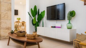 A modern living room with a wooden table, a white cabinet, a flat-screen TV, and large green plants. - Holiday Home "Ca Na Lola" Near the Beach with Wi-Fi, Air Conditioning & Terrace (Portocristo)