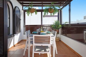 Un espace extérieur abrité, aménagé pour les repas, avec une table dressée pour quatre personnes, entourée de chaises blanches, d’une plante en pot et de jardinières suspendues.