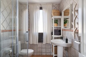 Une salle de bain équipée de toilettes, d’une douche et d’un lavabo, avec un carrelage au sol et un meuble blanc agrémenté de paniers.