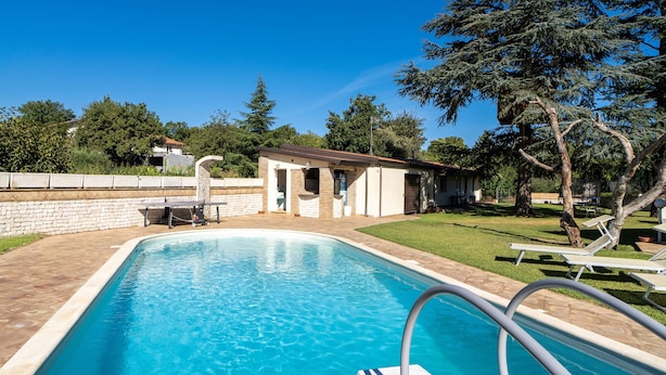 Une piscine à l’eau d’un bleu limpide, entourée d’une terrasse en briques, d’un petit bâtiment et d’un espace verdoyant planté d’arbres.