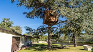 Une maison avec une cabane dans les arbres adossée à un grand arbre à feuilles persistantes, une terrasse avec des meubles d’extérieur et un garage.