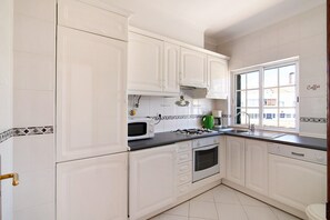 Une cuisine avec des meubles blancs, un four à micro-ondes, une cuisinière à gaz et une fenêtre donnant sur des immeubles.
