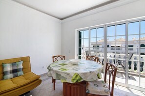Un coin repas avec une table dressée pour quatre personnes, un canapé agrémenté d’un coussin à carreaux et une grande fenêtre donnant sur des immeubles.