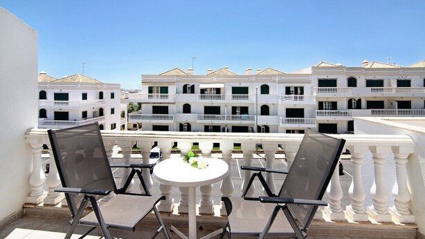 Un balcon avec deux chaises et une table, donnant sur une rangée de bâtiments blancs, eux aussi dotés de balcons.