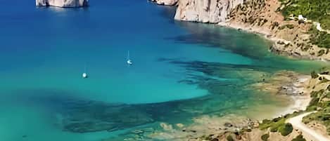 A coastal landscape with clear waters, rocky cliffs, and a beach with a path leading to a building.