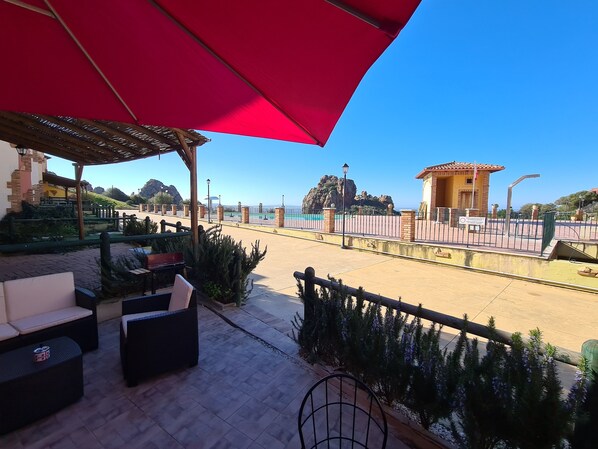 Une terrasse avec des sièges extérieurs, un parasol rouge et une vue sur la plage et le paysage rocheux.