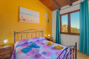 A bedroom with a bed, a nightstand, a lamp, a painting, and a window with curtains.