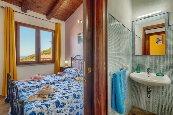 A bedroom with two beds, a window, a bathroom with a sink and mirror, and a view of the outdoors.
