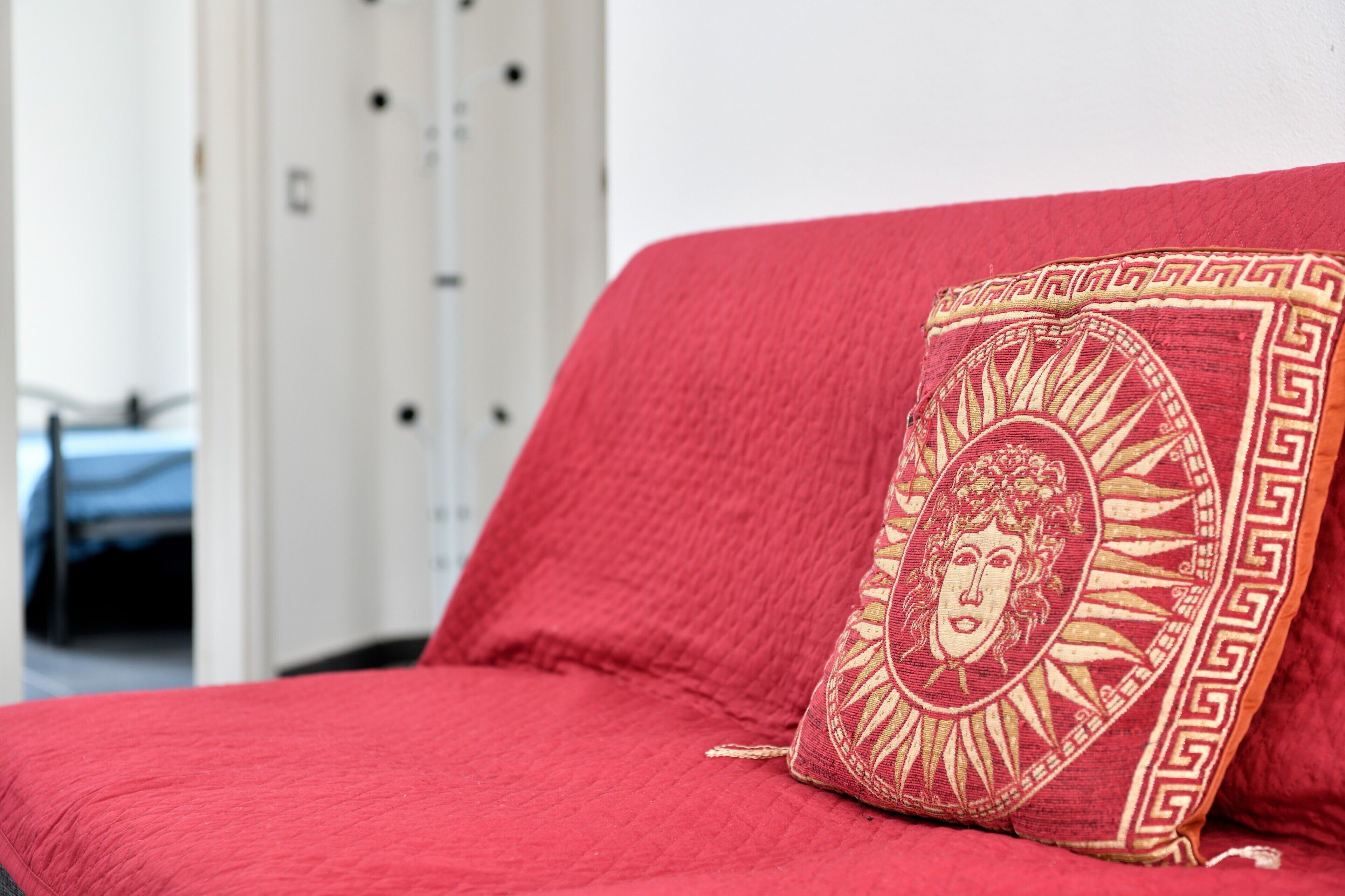 Un sofà vermell amb un coixí decoratiu en una habitació amb un armari blanc i un llit al fons.