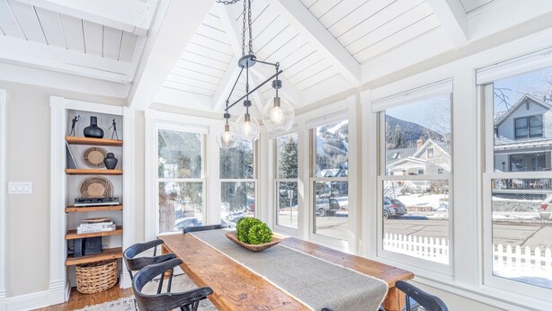 A dining area with a wooden table, chairs, a chandelier, and large windows with a view of houses and a snowy street. - Sunnyside Retreat Home (Telluride)