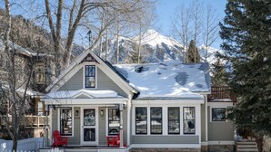 A two-story house with a snow-covered roof, a porch with red chairs, and a white picket fence. - Sunnyside Retreat Home (Telluride)